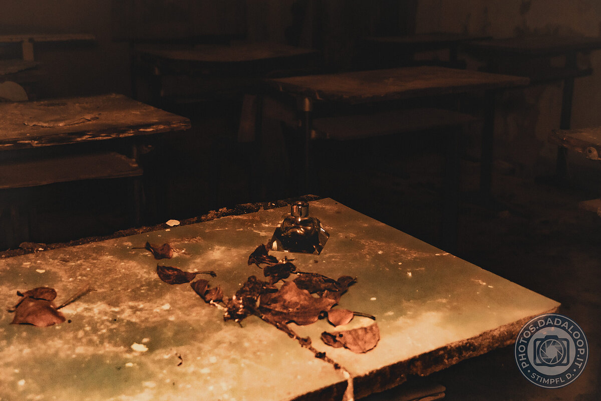 Dettaglio di un banco polveroso con calamaio e foglie in una scuola abbandonata